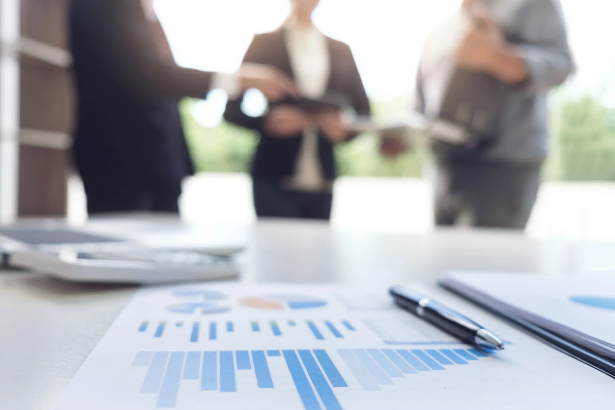 Des documents avec graphiques posés sur une table, des collègues discutent en arrière-plan