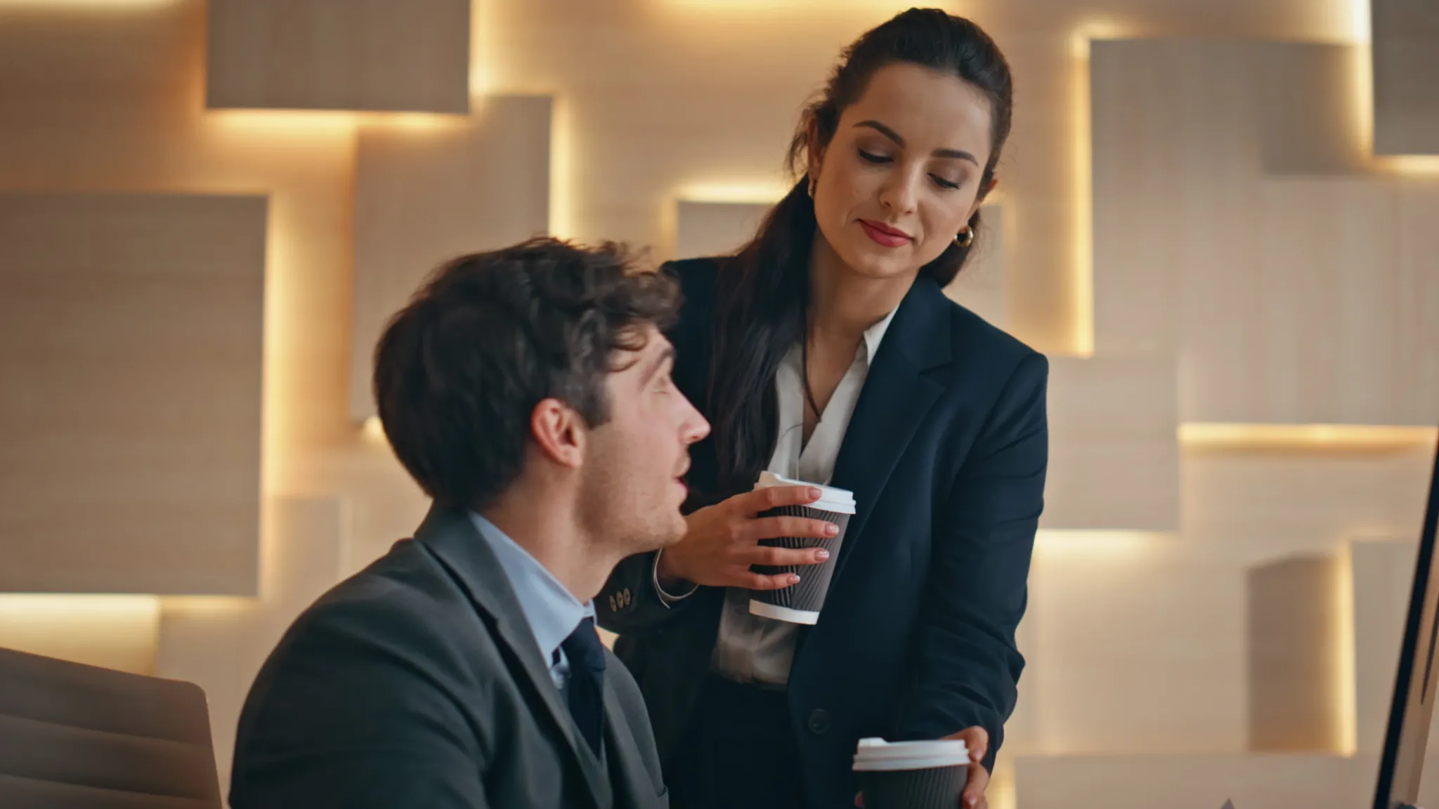 Une femme, un gobelet de café à la main, discute avec un collègue assis à son bureau