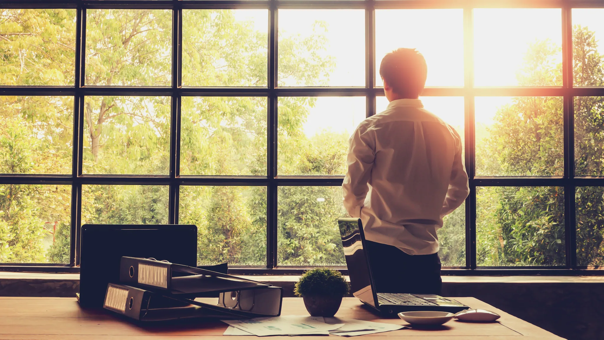 Un homme se tient debout face à une grande fenêtre ouverte sur la verdure, son bureau derrière lui